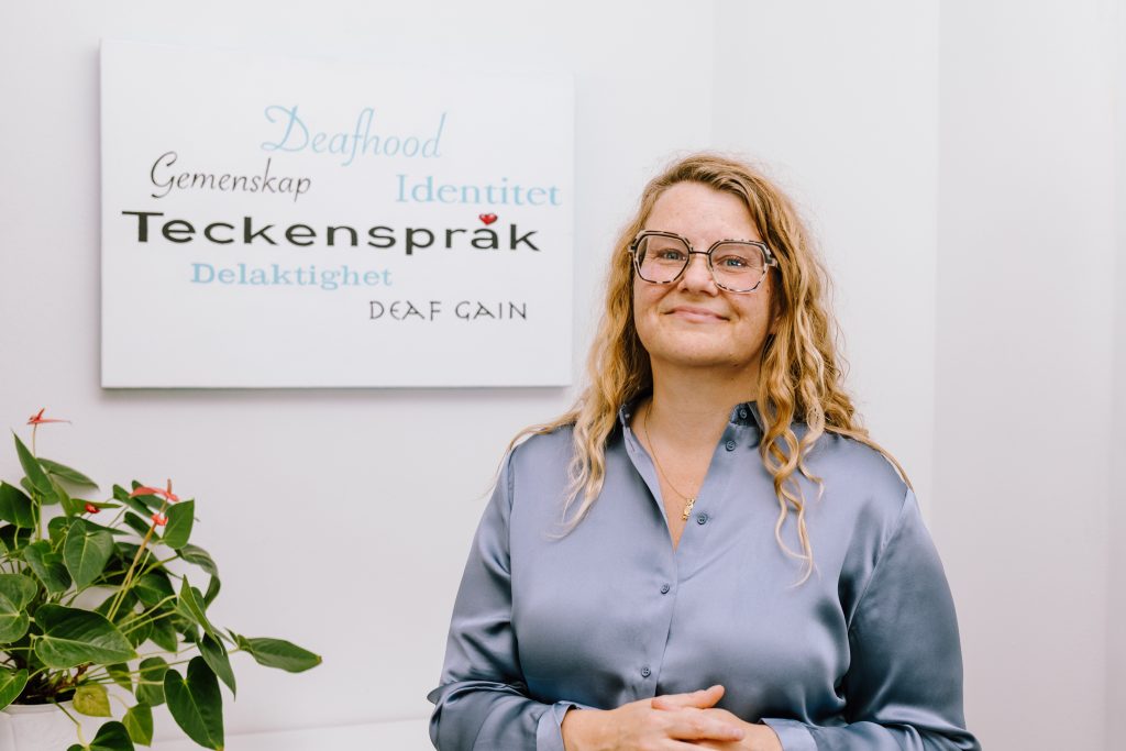 Hedvig Ledung. Bakom henne finns en tavla på väggen där det står bland annat teckenspråk, deafhood och identitet.